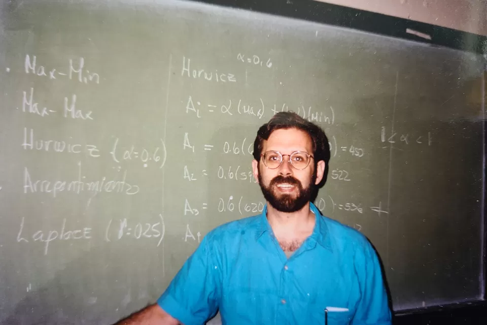 Photo of Bruce Stiftel as a young man standing in front of a green chalkboard